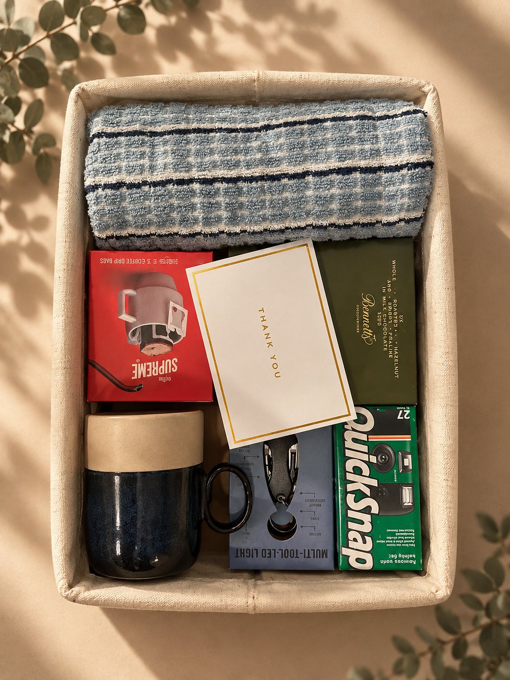An open gift basket holding a striped towel, coffee drip bags, chocolate, a stoneware mug, a multi tool and a film camera, with a gold edged thank you card