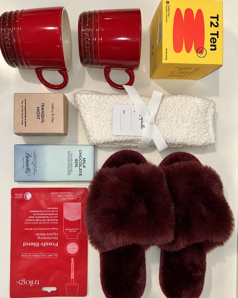 A gift of two red mugs, tea, a candle, chocolate, a face mask, socks and burgundy slippers